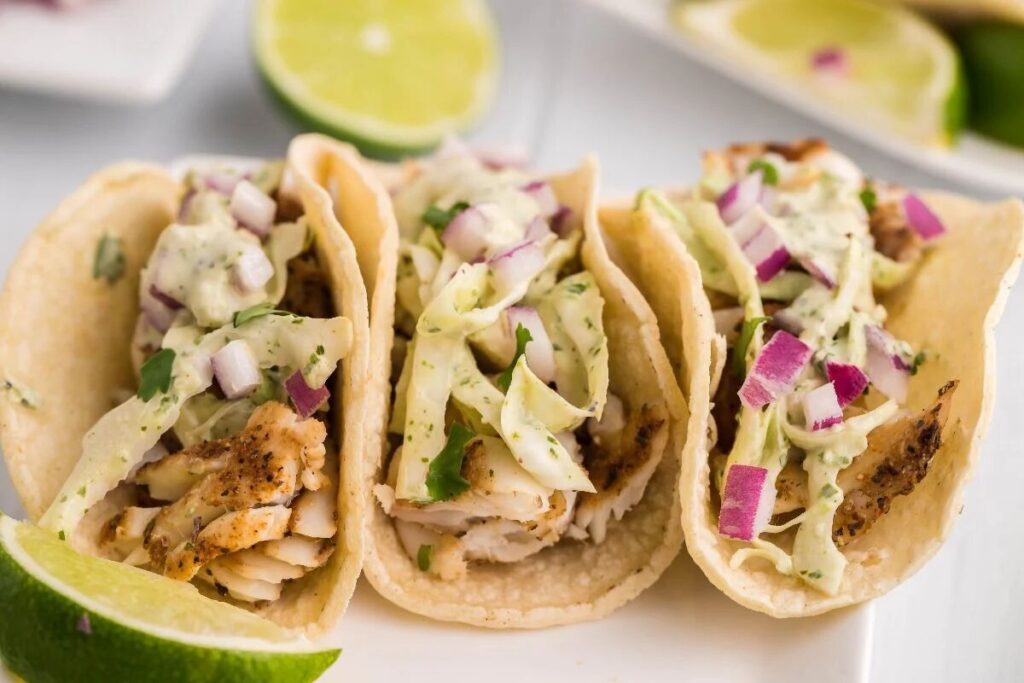 Three smoked fish tacos with cabbage slaw for fish tacos, diced red onion, and creamy sauce, served with fresh lime wedges.