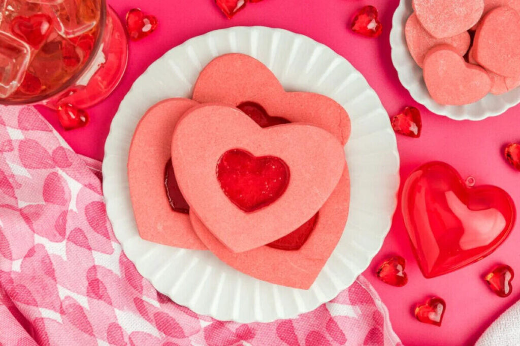 Heart-shaped pink stained glass cookies with candy centers and cutout details, arranged on a white plate with heart decorations.