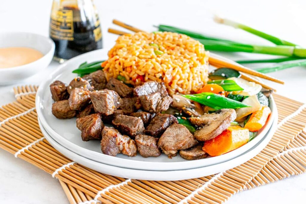 Hibachi steak cubes with fried rice and sautéed vegetables, served with chopsticks and soy sauce.