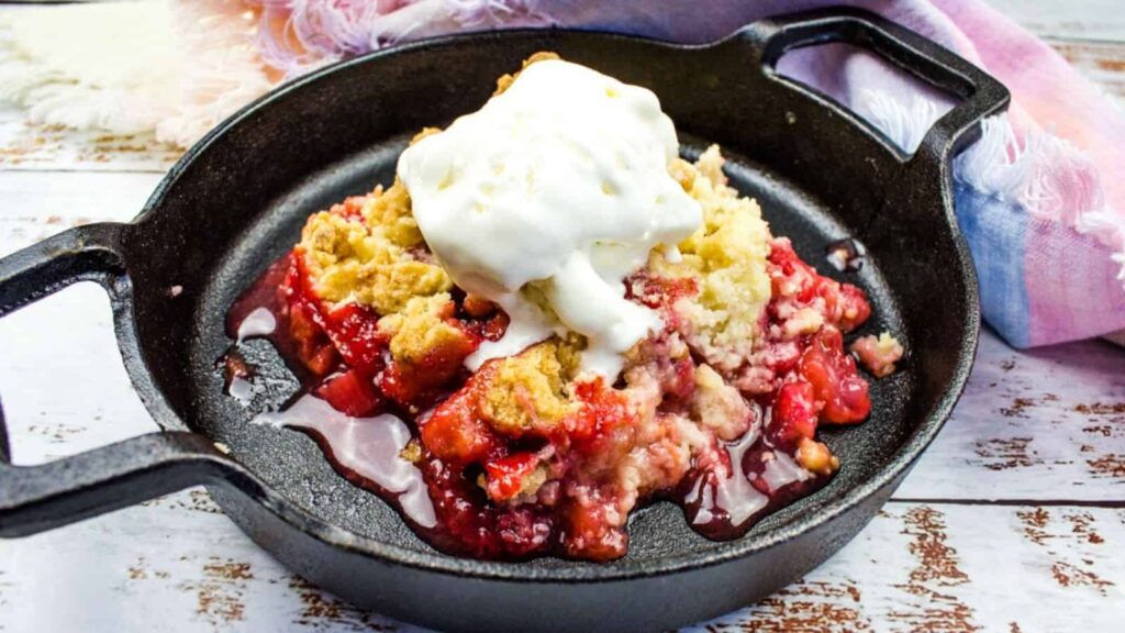 Warm strawberry crumble topped with vanilla ice cream, served in a black cast iron skillet, with a colorful cloth in the background, set on a distressed white-painted wooden table.