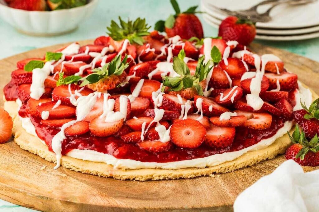 A strawberry pizza topped with sliced strawberries and drizzled with white icing rests on a wooden board, while plates and utensils linger in the background.