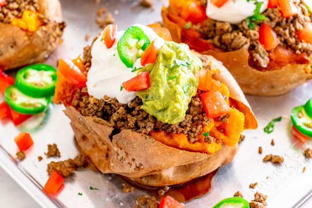 Loaded sweet potato with spiced meat, guacamole, sour cream, tomato, and jalapeños on a tray.