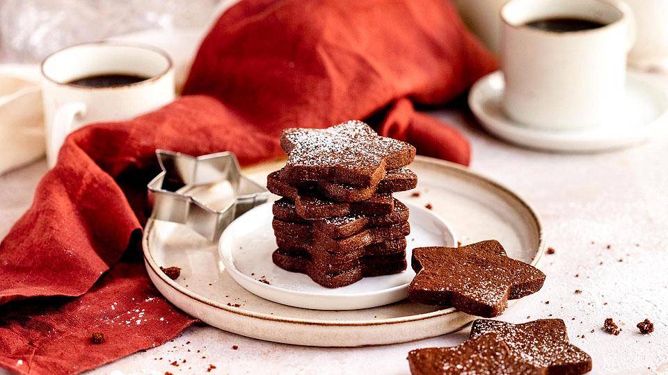 Star-shaped chocolate cut-out cookies stacked on a small plate with a metal star cookie cutter and two cups of coffee visible nearby.