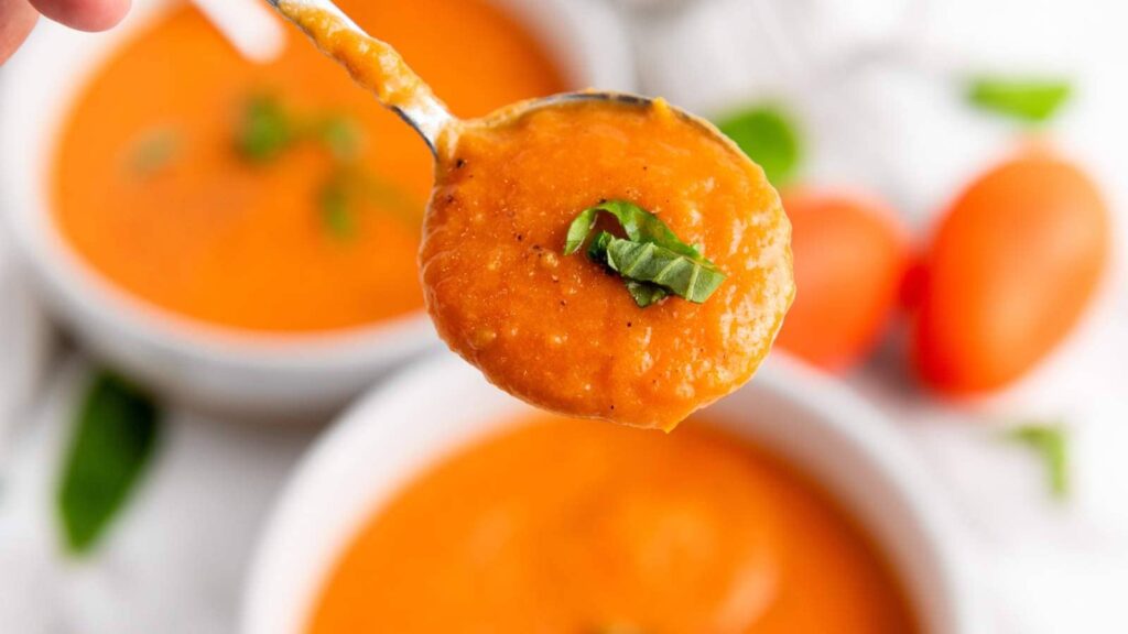 A spoonful of orange soup garnished with green herbs, with bowls of the same soup in the background.