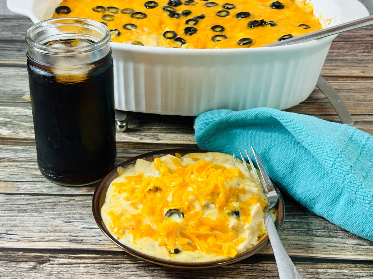 A plate of creamy casserole topped with melted cheddar cheese and black olive slices sits beside a jar of dark soda. A larger serving dish of the same casserole is in the background on a wooden table. A fork rests on the plate.