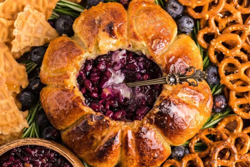 Round baked brie cheese dip with berry filling, surrounded by pretzels, waffle cookies, and fresh blueberries for a delightful blueberry brie appetizer.
