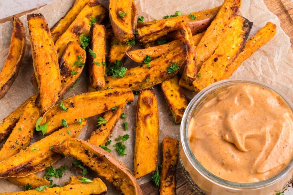 Air fryer sweet potato wedges garnished with chopped herbs lie next to a glass container of dipping sauce on parchment paper.