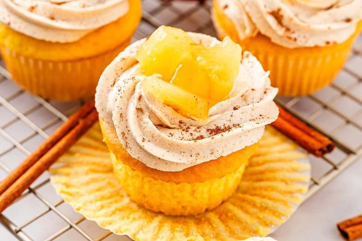 Close-up of cupcakes topped with frosting and apple chunks, sprinkled with cinnamon. A knife is spreading the frosting.
