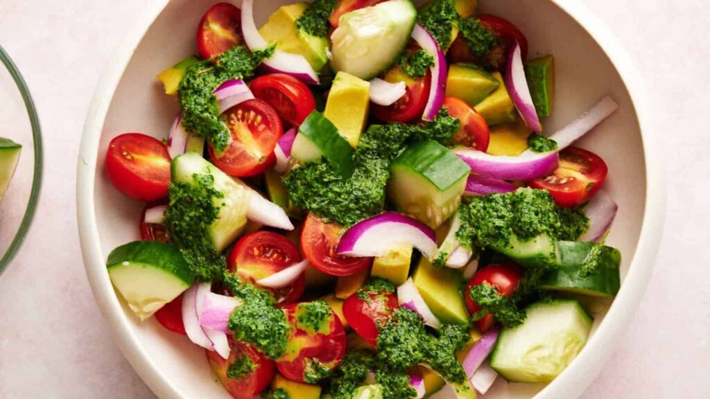 Overhead shot of a bowl of fresh salad with halved red cherry tomatoes, sliced cucumbers and red onions, avocado pieces, and a vibrant green pesto dressing.