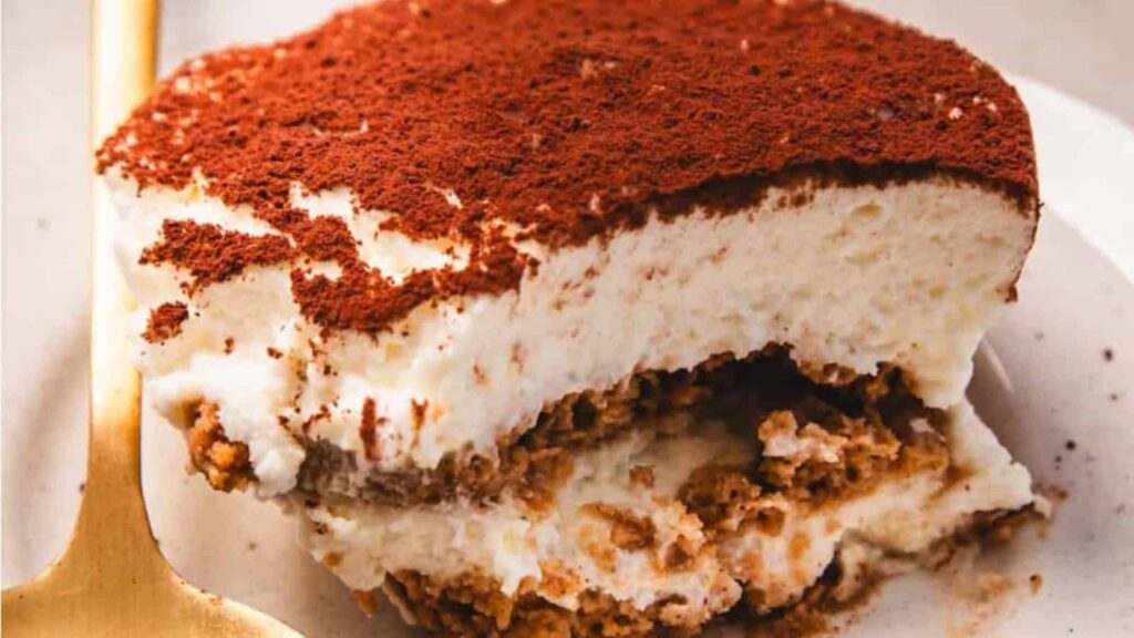 A slice of tiramisu dessert topped with cocoa powder on a plate with a gold-colored fork.
