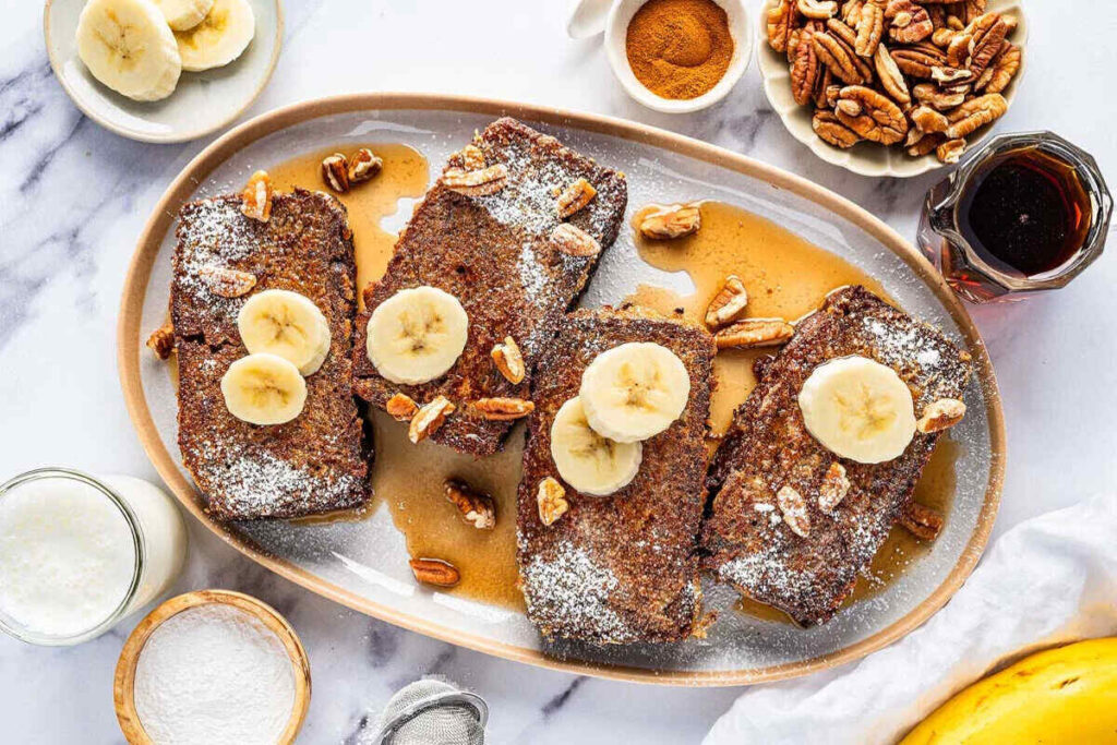 A plate of banana bread French toast slices with powdered sugar, pecans, and syrup is surrounded by ingredients like bananas, pecans, cinnamon, and a glass of milk on a marble surface.