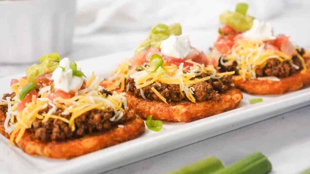 Three hash brown patties topped with ground beef, cheese, sour cream, tomatoes, and green onions on a plate.