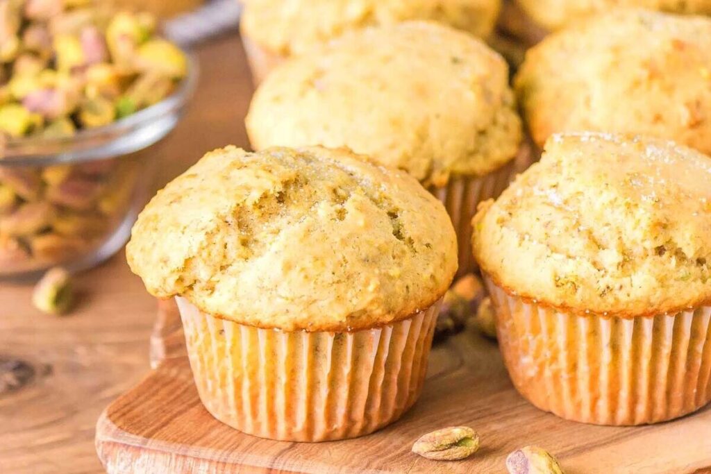 A close-up of several freshly baked pistachio muffins on a wooden surface, with a bowl of whole pistachios in the background.