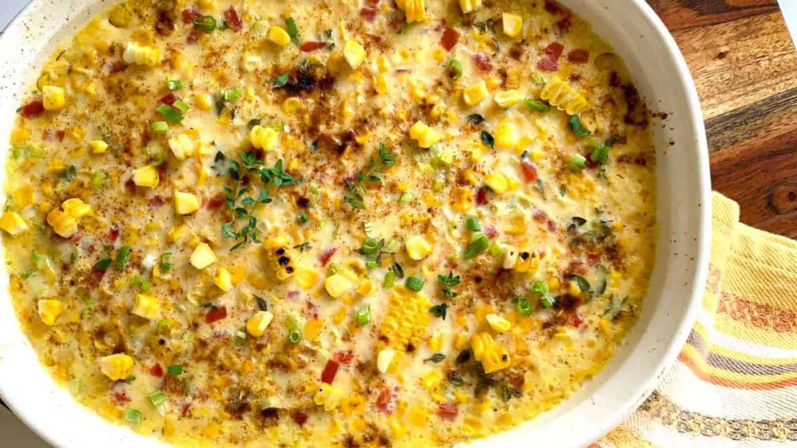 A baked corn casserole with chopped peppers and herbs is presented in a white oval dish on a wooden surface, with a patterned cloth nearby.