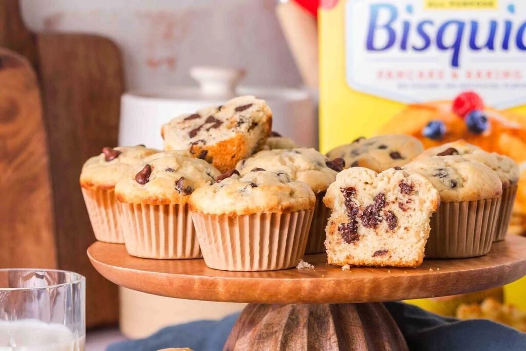 Bisquick chocolate chip muffins on a wooden stand, with one muffin cut in half showing the inside goodness. A box of Bisquick mix is visible in the background, promising homemade perfection.