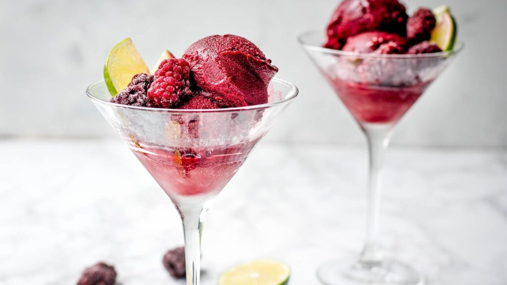 Horizontal image of blackberry lime sorbet in a metal loaf pan on a marble background garnished with frozen blackberries and limes.