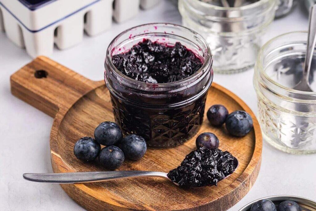 A jar of blueberry jam on a wooden board, surrounded by fresh blueberries and a spoonful of rich blueberry jam.