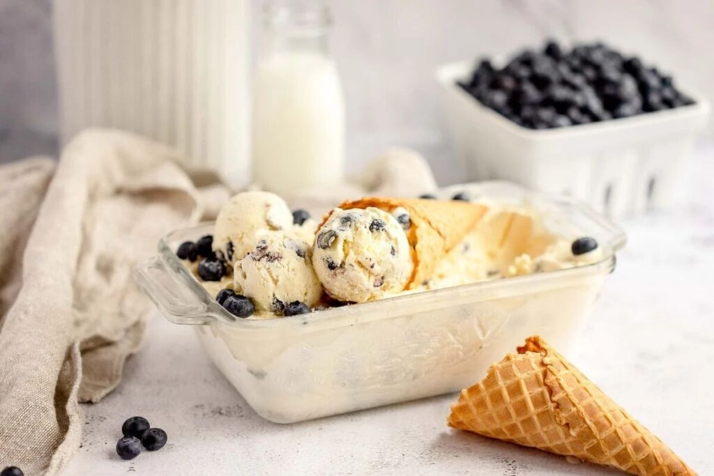 Glass dish with Blueberry Cheesecake Ice Cream, a waffle cone, fresh blueberries, and a bottle of milk in the background.