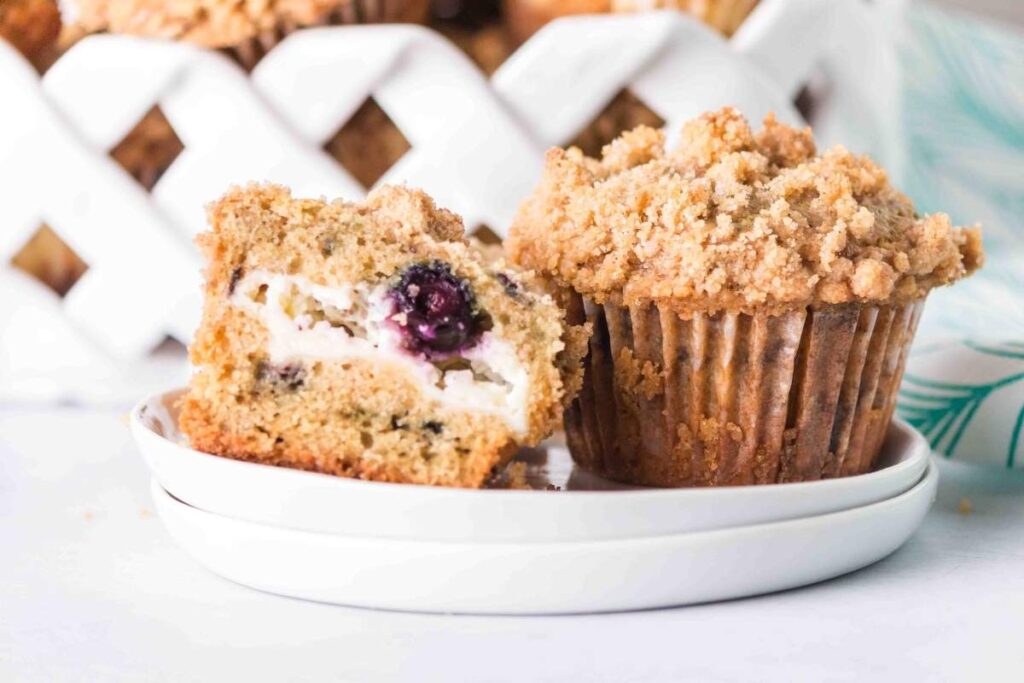A blueberry cream cheese muffin topped with crumbly streusel sits on a plate next to a muffin cut in half, revealing its lush filling. In the background, a basket brims with more of these delectable treats.
