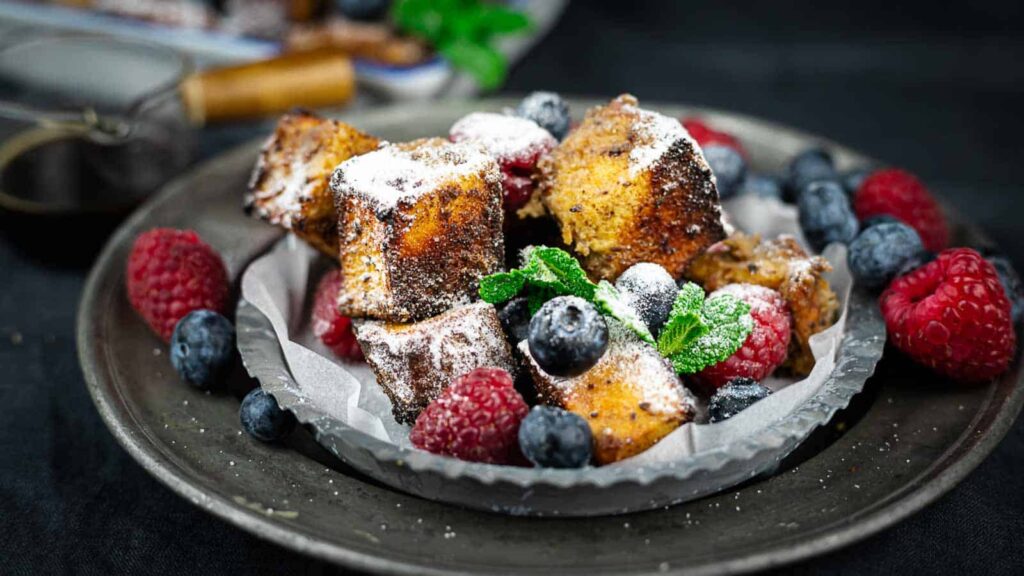 Close-up shot of a plate of French toast casserole, dusted with powdered sugar and garnished with fresh raspberries, blueberries, and mint leaves, arranged on a rustic, pewter-colored plate.