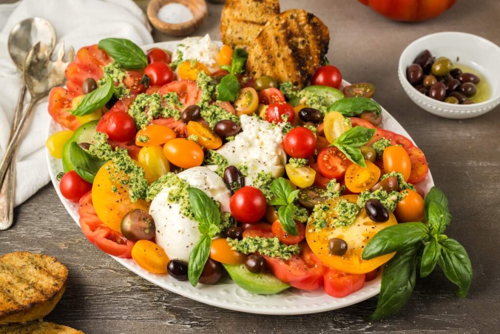 A burrata with tomatoes and pesto appetizer or salad featuring fresh basil and olives, all beautifully arranged on a white platter, accompanied by grilled bread slices on the side.