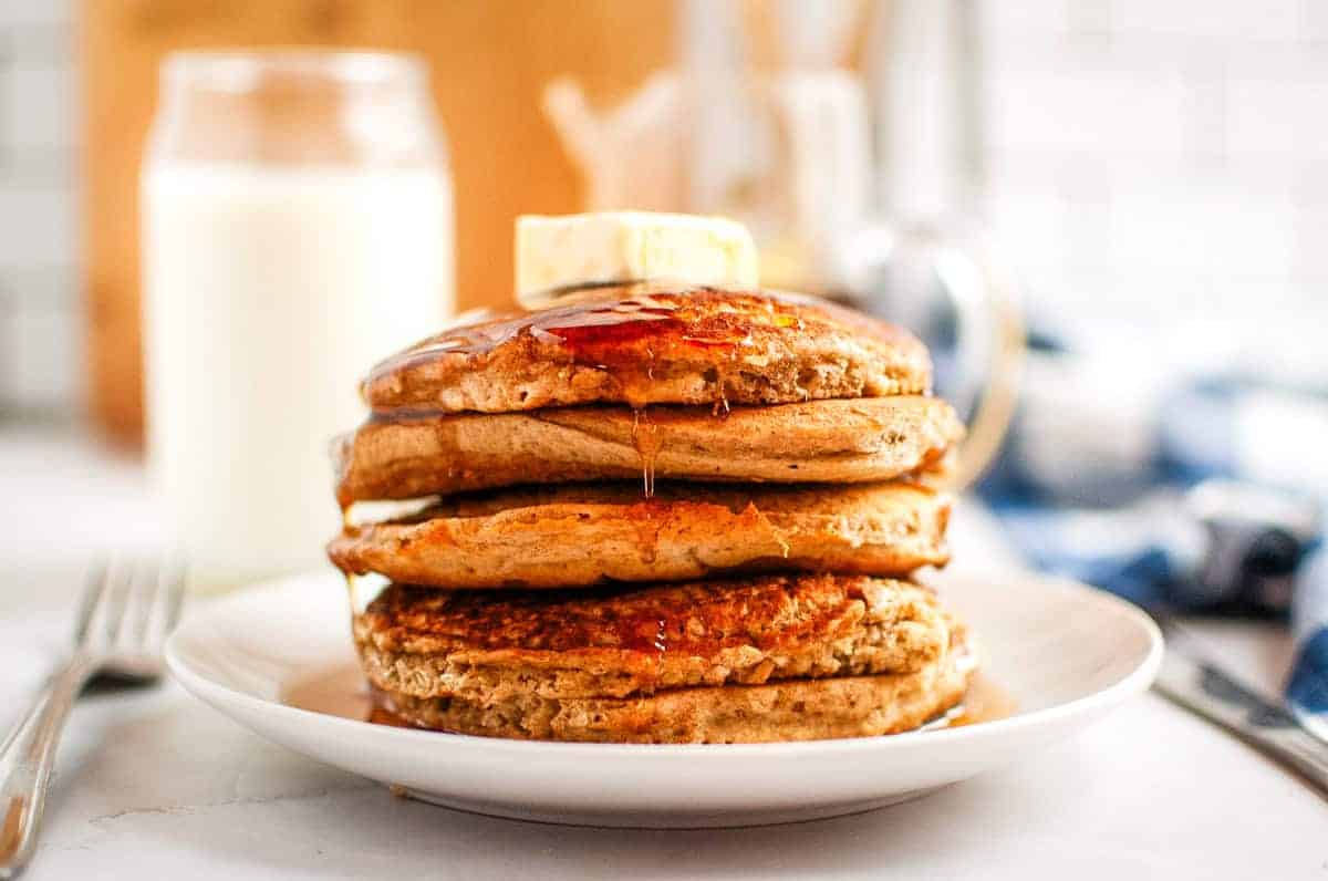 A stack of pancakes with a pat of butter on top is drizzled with syrup on a white plate. A glass of milk and a striped napkin are in the background.