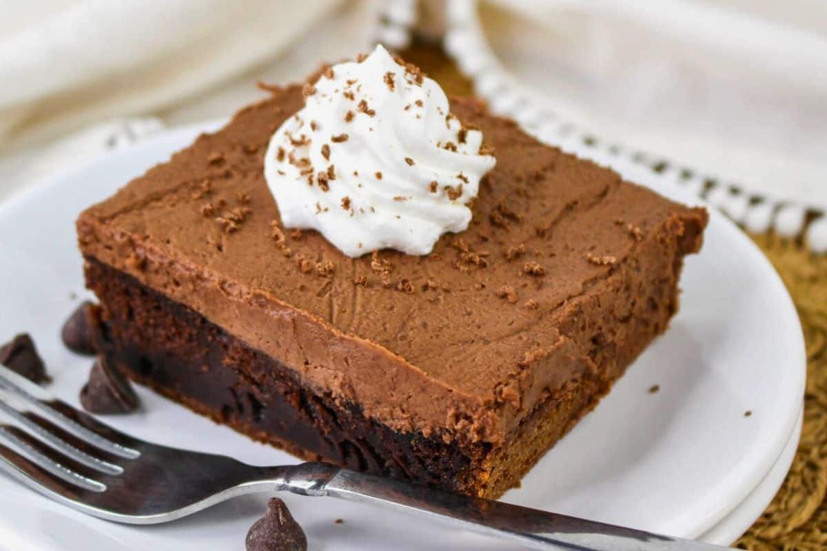 A slice of layered chocolate dessert with whipped cream on top, served on a white plate with a fork and chocolate chips nearby.