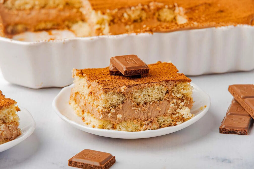A slice of chocolate tiramisu topped with cocoa powder and a square of chocolate on a white plate, with a fork nearby.