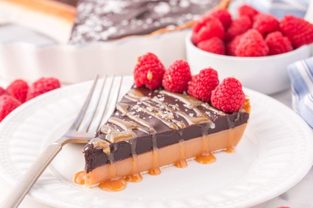A slice of rich chocolate caramel tart topped with fresh raspberries sits on a white plate, accompanied by a fork. In the background, a bowl brims with juicy raspberries.