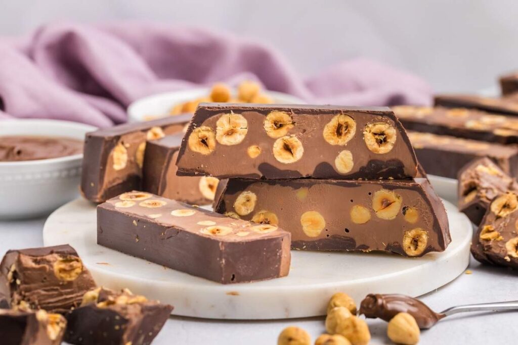 Chocolate hazelnut torrone bars with whole hazelnuts rest elegantly on a marble plate, surrounded by scattered hazelnuts and a bowl of rich chocolate spread.