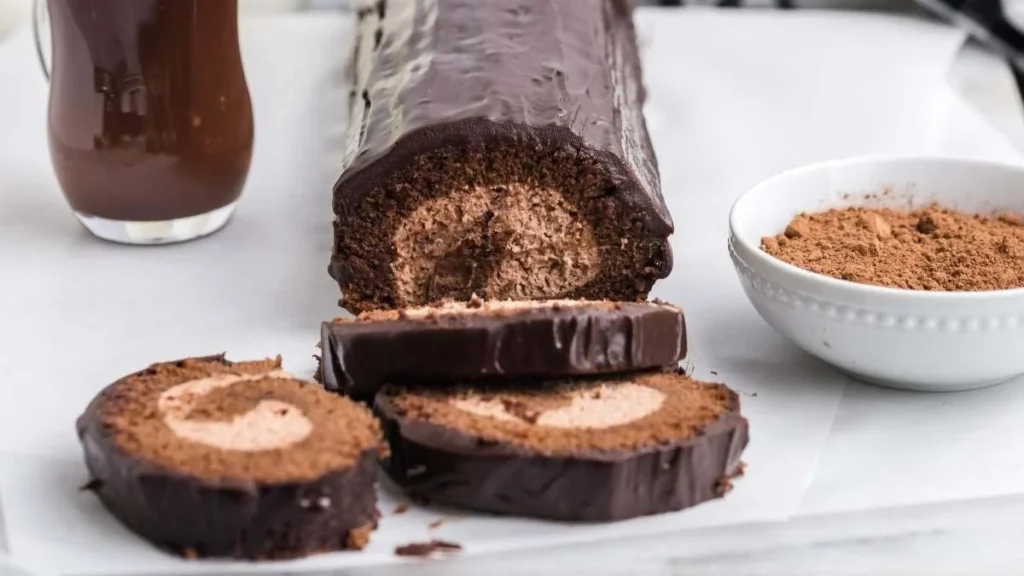 Chocolate swiss roll with three slices cut, revealing cream filling. A small bowl of cocoa and a jug of chocolate sauce are beside it on a white surface.