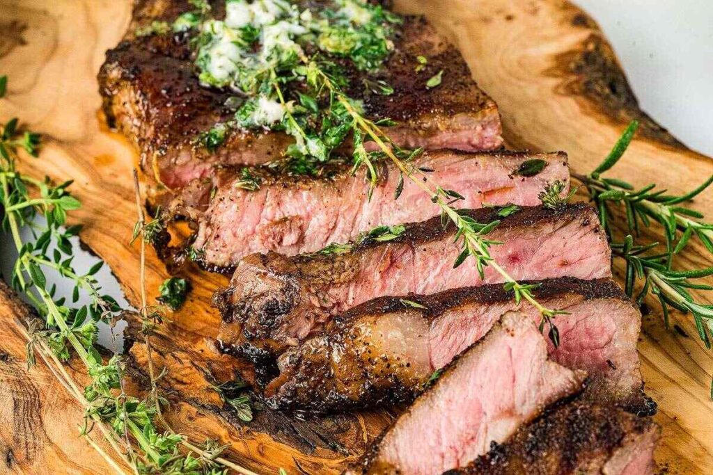 A sliced medium-rare coffee-rubbed steak garnished with herbs and a dollop of herb butter, served on a wooden cutting board.