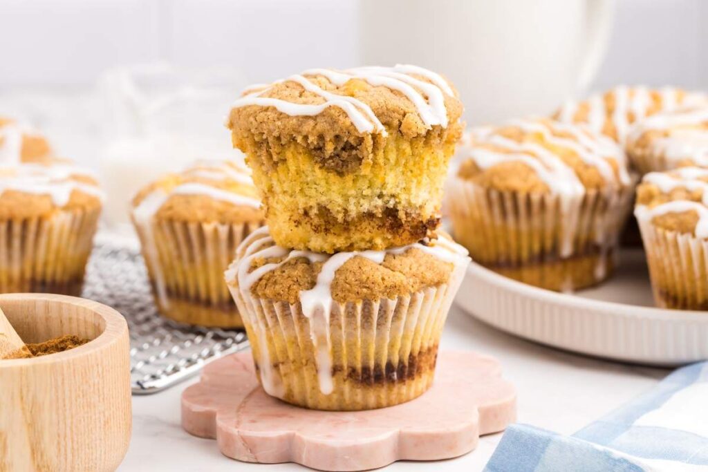 Two stacked coffee cake muffins with icing drizzle on a small plate; several more muffins are on a wire rack and a plate in the background.