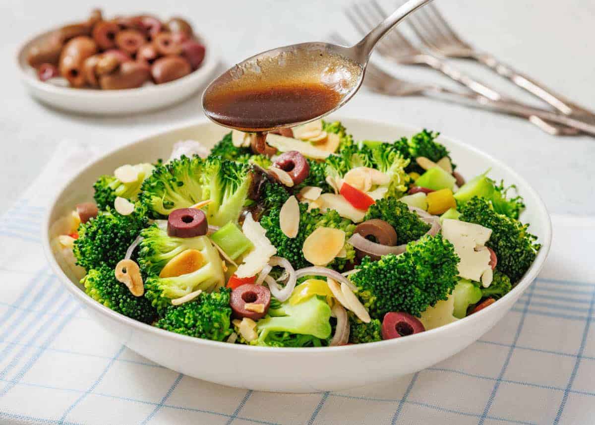 A bowl of Italian broccoli salad with sliced olives, almonds, onions, and red peppers, served on a white plate with a fork and napkin beside it.
