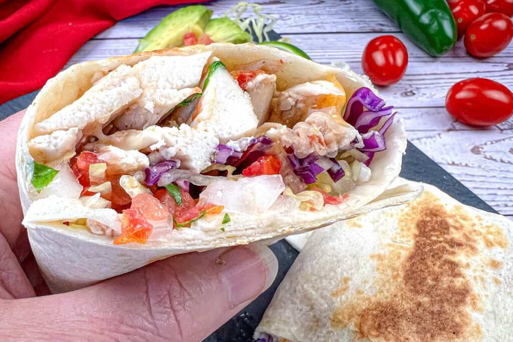 Hand-held open burrito filled with large pieces of white chicken, diced red tomatoes, red cabbage, sauce, and lettuce, next to a partially visible burrito.