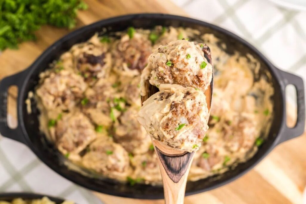 A wooden spoon holds creamy meatballs above a dish filled with more meatballs in a luscious sauce.