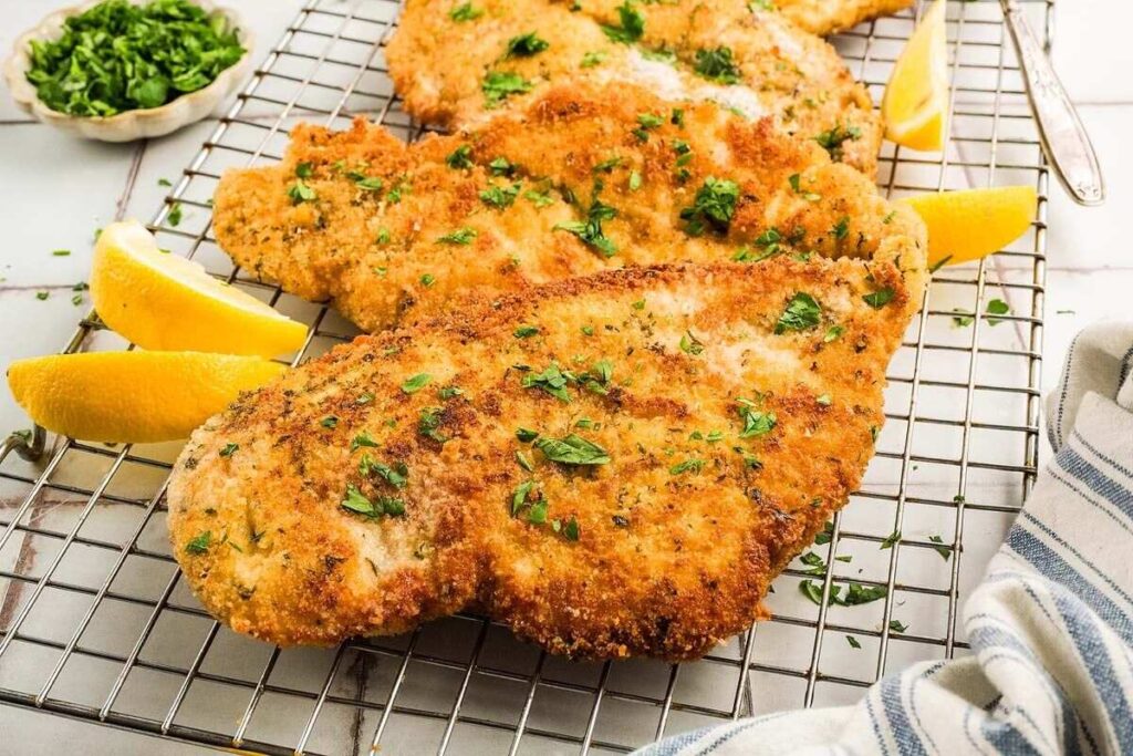 Crispy chicken cutlets garnished with parsley rest on a wire rack, accompanied by lemon wedges and a small bowl of fresh herbs.