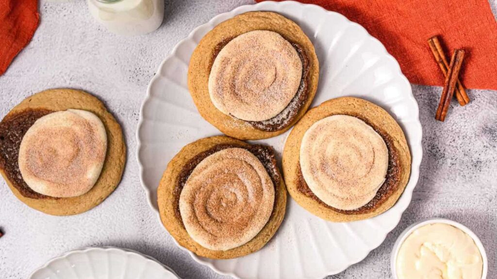 Four cinnamon swirl cookies on a white plate with a glass of milk and cinnamon sticks nearby.