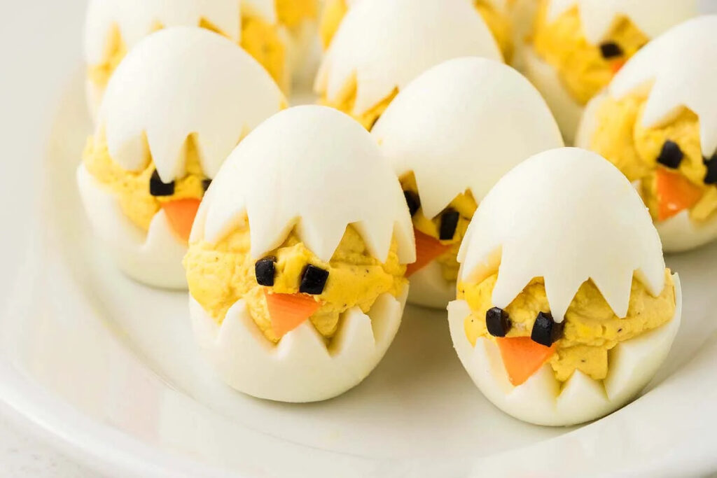 Deviled egg chicks adorned with olive eyes and carrot beaks perch adorably on a white plate.