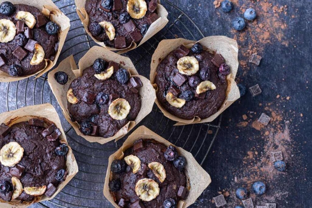 Six Double Chocolate Blueberry Banana Muffins topped with banana chips and blueberries on a wire rack, with cocoa and blueberries scattered nearby.