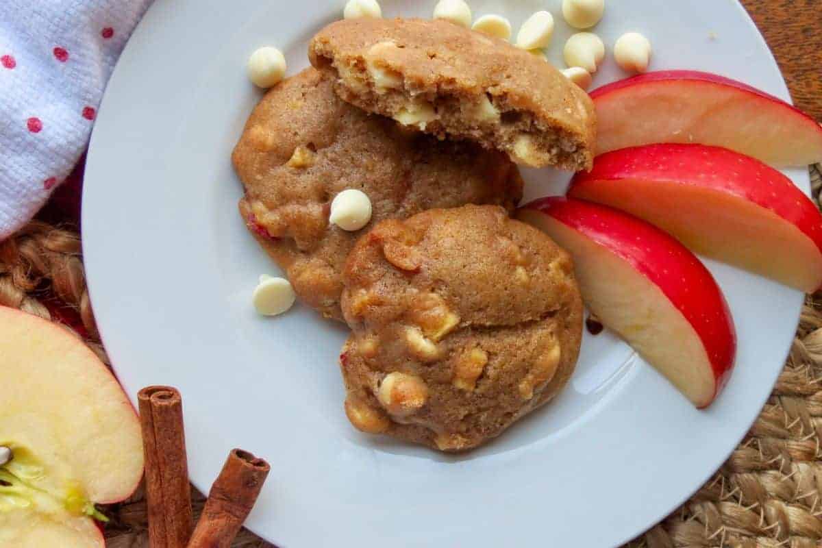 A plate with applesauce cookies containing white chocolate chips, garnished with apple slices and surrounded by cinnamon sticks and a red apple.
