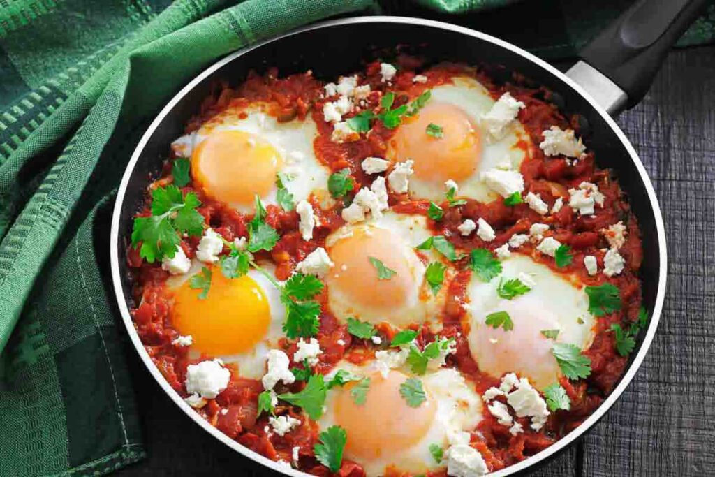 A skillet filled with eggs cooked in tomato sauce, topped with crumbled cheese and fresh parsley, sits next to a green cloth.
