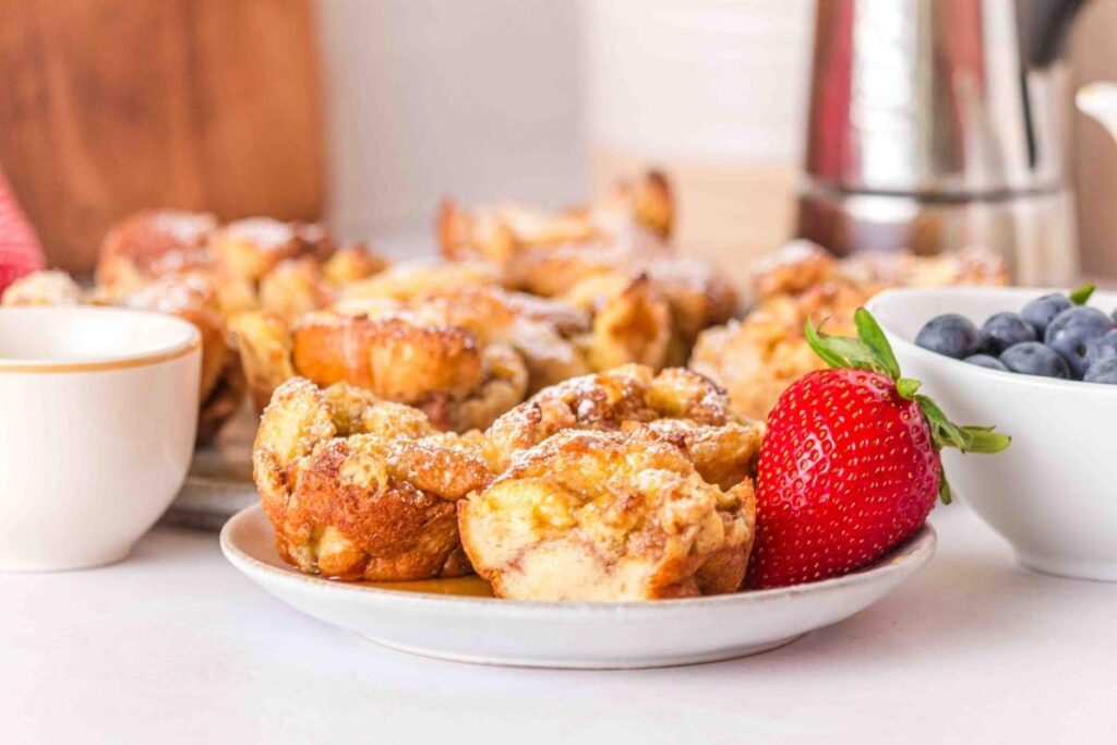 A plate of French toast muffins topped with powdered sugar, accompanied by a strawberry and a bowl of blueberries, with more pastries and a coffee pot in the background.