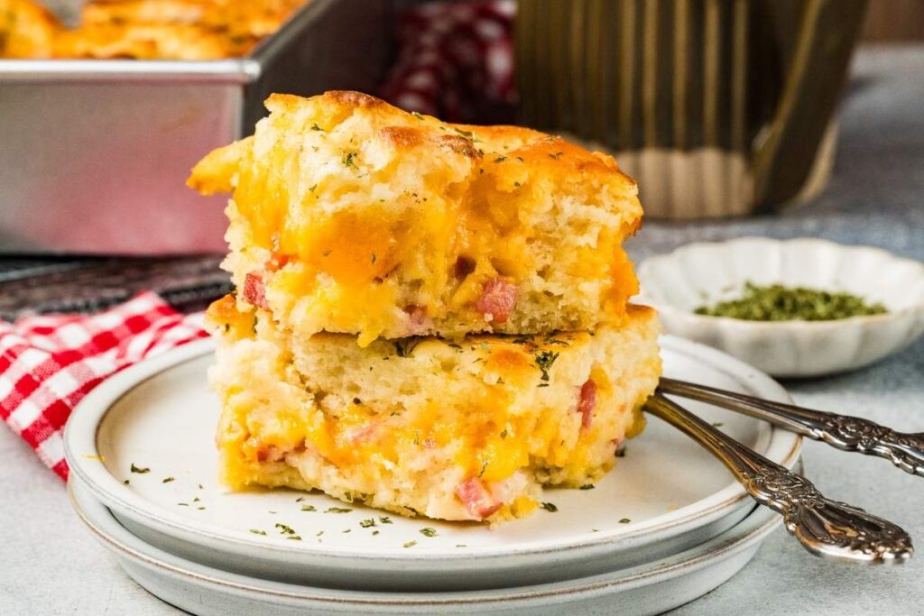 Two slices of ham and cheese butter swim biscuits on a white plate, silverware beside them, baking dish in the background.
