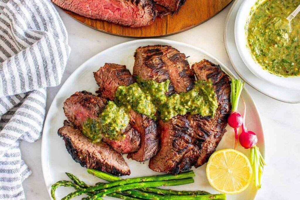 Plate of Italian steak slices topped with green salsa verde sauce, accompanied by asparagus, radishes, and a lemon half. A bowl of extra sauce is on the side.