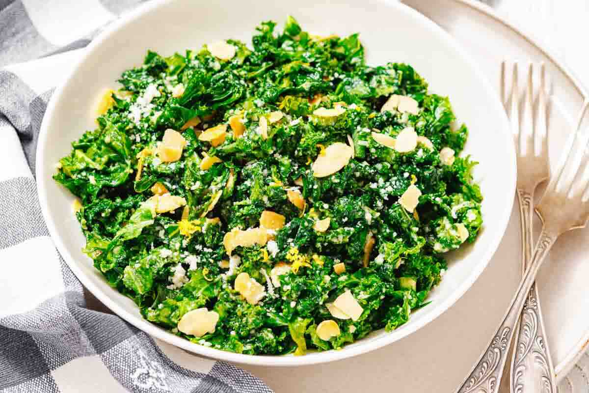 A white bowl filled with chopped kale salad topped with sliced almonds and grated cheese, placed on a plate beside two forks and a checkered napkin.