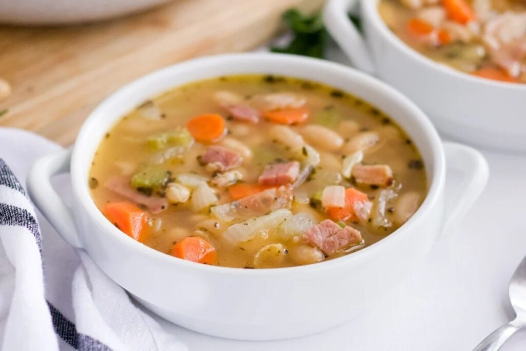 A bowl of leftover ham and cannellini bean soup features white beans, sliced carrots, celery, and tender ham pieces infused with herbs in a savory broth.