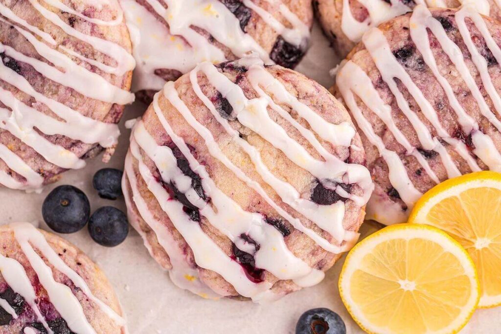 Lemon blueberry cookies adorned with a zesty lemon icing, nestled amidst fresh blueberries and vibrant lemon slices on a light-colored surface.