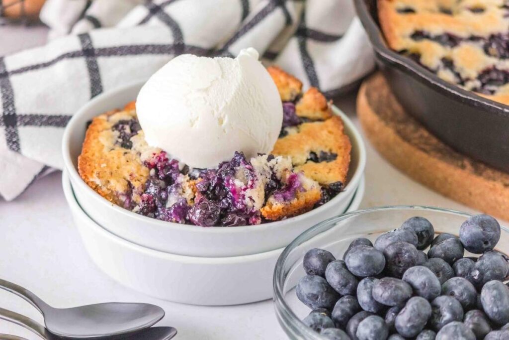 A bowl of Bisquick lemon blueberry cobbler with a scoop of vanilla ice cream on top sits beside a bowl of fresh blueberries, with a cast iron skillet and checkered towel in the background.