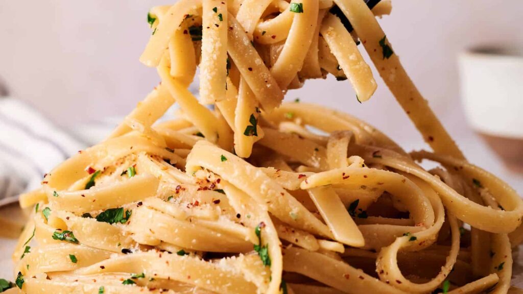 Close-up of a fork twirling fettuccine pasta topped with grated cheese, chopped parsley, and red pepper flakes.